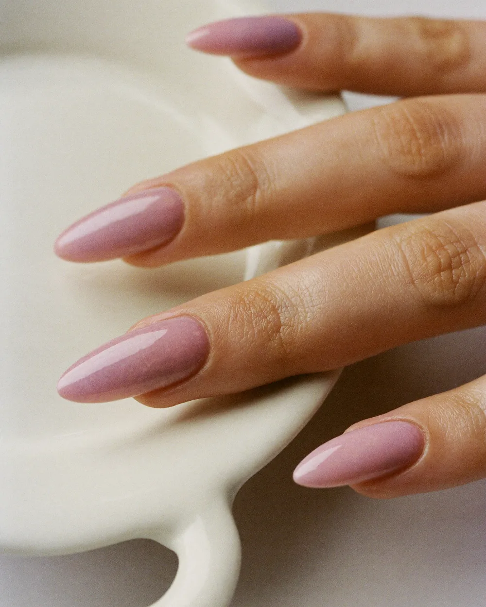 A hand resting on the rim of a cream ceramic dish, long almond nails in a glossy milky dusty rose.
