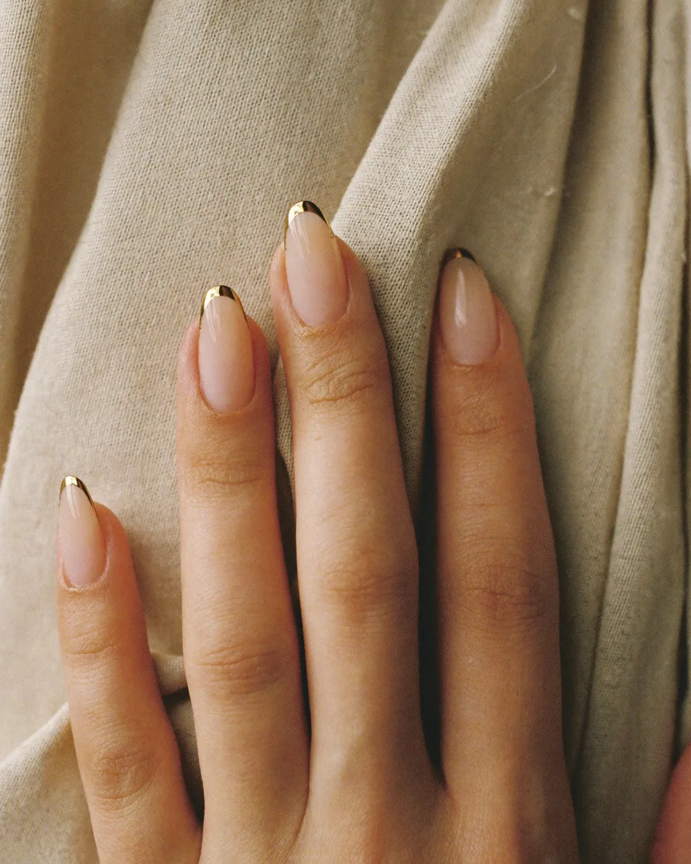 Long almond nails on a sheer nude base, each finished with a mirror-gold French tip, against oat-coloured linen.