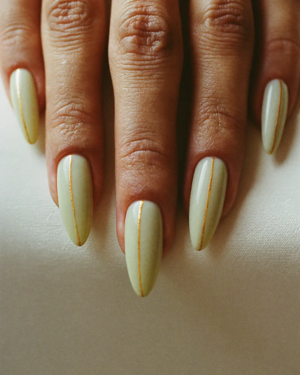 Long almond nails in a pale pistachio cream, each with one fine gold line hand-painted down the length of the nail.