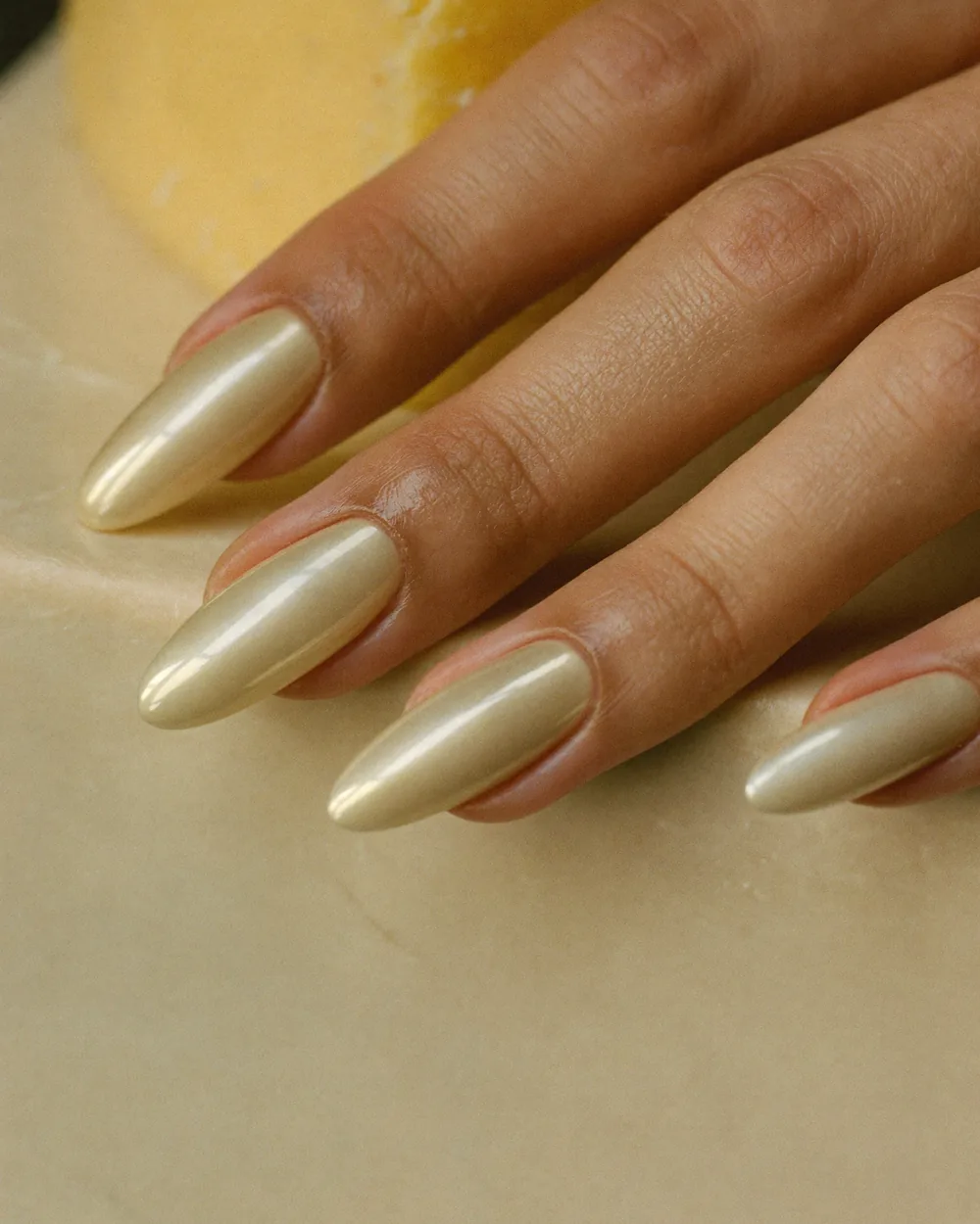 Long almond nails in a solid pearl-cream chrome with no tip, on a hand resting against a pale cream surface.