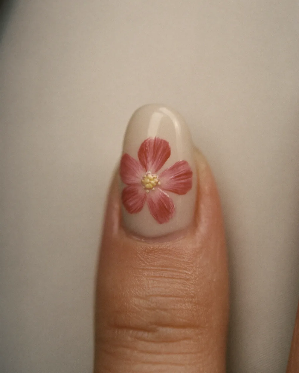 A close crop of a single thumb, the short milky-cream nail carrying one hand-painted dusty-rose flower with a small gold centre.